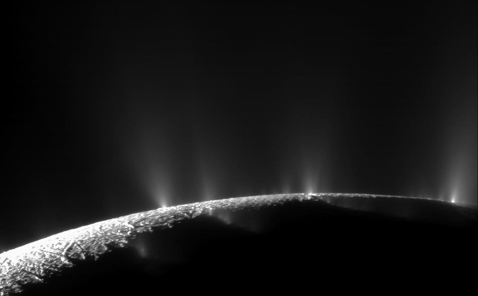 Enceladus backlit by the Sun, showing bright plumes of water vapor and ice particles jetting from its south pole against black space.