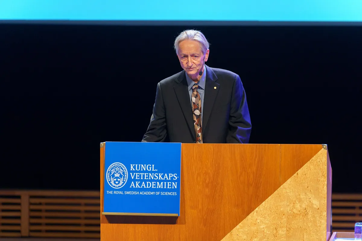Geoffrey Hinton standing at a wooden lectern bearing the Royal Swedish Academy of Sciences sign, delivering a lecture against a dark stage.