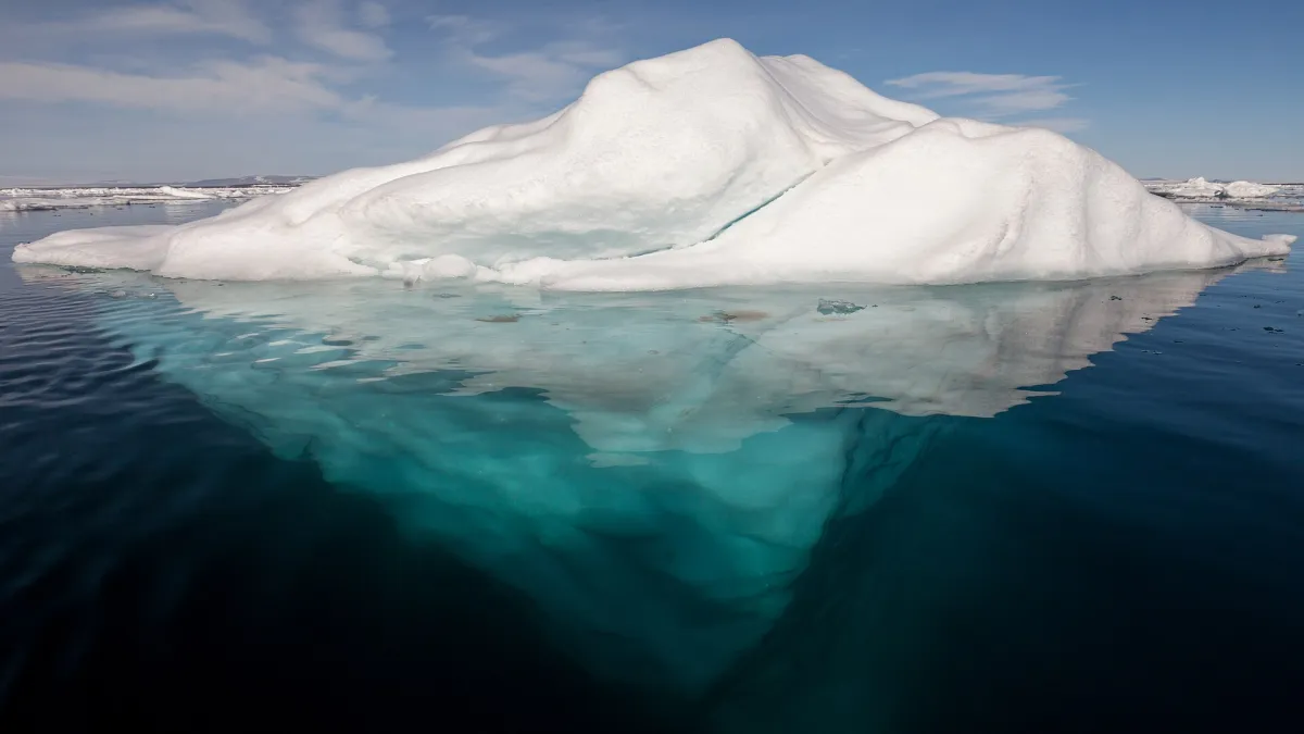 An iceberg in the Arctic photographed at the waterline: a small white crown above the surface, and a far larger turquoise mass descending into dark water below.